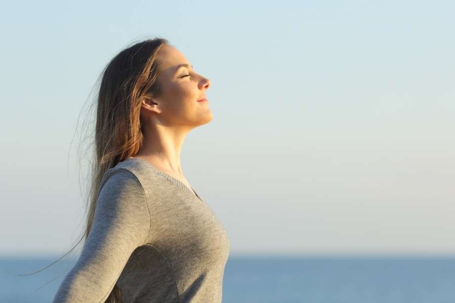 Une femme respire profondément les yeux fermés en plein air face à la mer pour décompresser.