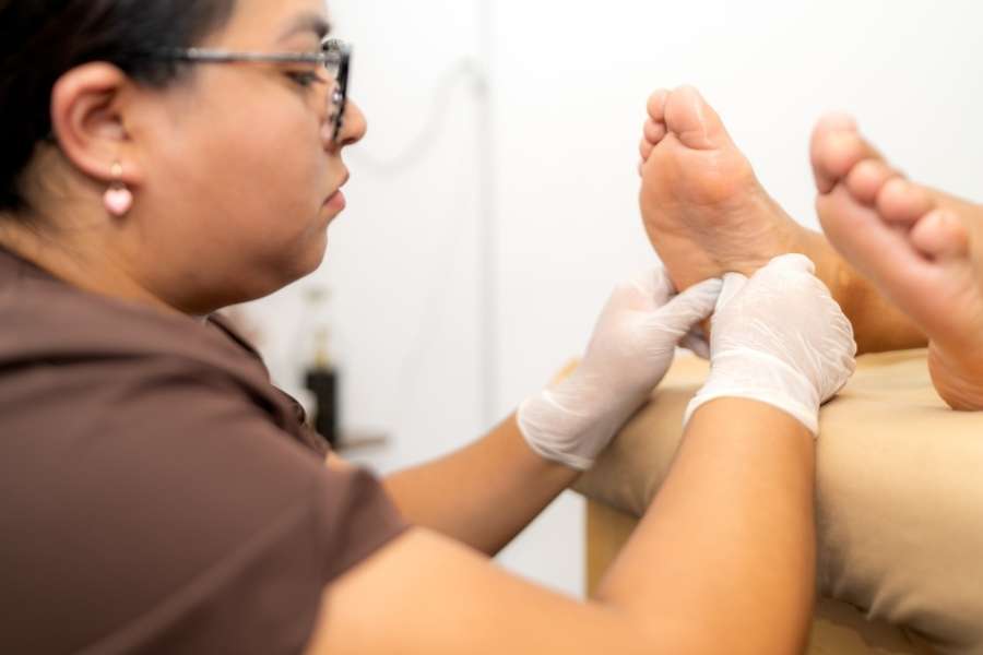 Une praticienne en blouse marron et gants blancs effectue un massage des pieds en réflexologie plantaire.
