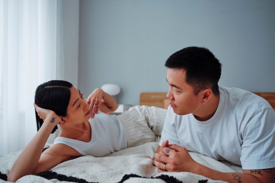 Un couple complice allongé sur un lit blanc, se regardant avec tendresse pour illustrer une sexualité épanouie.